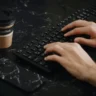 Dłonie na czarnej klawiaturze na ciemnym marmurowym biurku, obok papierowy kubek i telefon